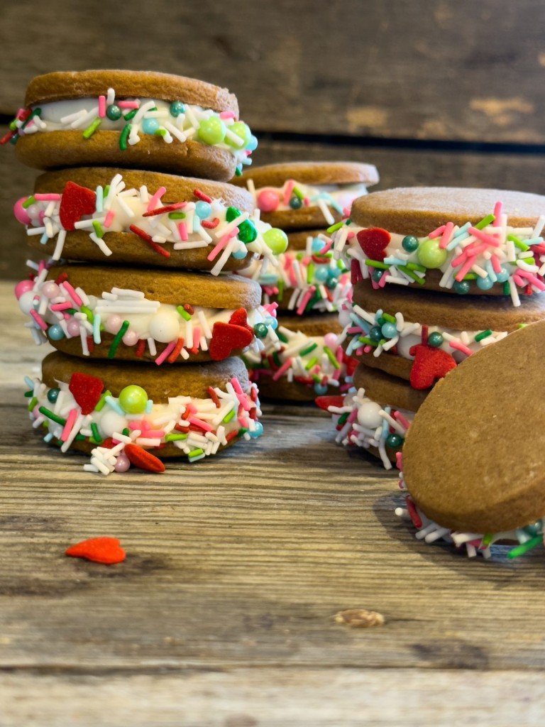 Close-up of gingerbread sandwich cookies made with Pillsbury gingerbread dough, filled with creamy vanilla frosting, and rolled in vibrant holiday sprinkles, displayed on a rustic wooden surface.