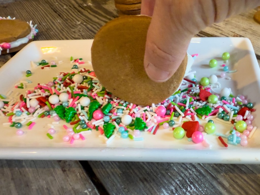Hand dipping a gingerbread sandwich cookie with frosting into colorful holiday sprinkles on a white plate for a festive dessert.