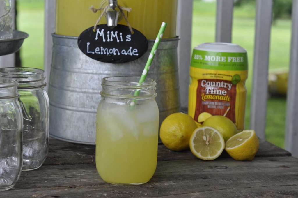 
A mason jar filled with MiMi’s Lemonade and a green polka dot straw sits on a rustic wooden table. Behind it, a galvanized drink dispenser labeled “MiMi’s Lemonade,” fresh lemons, and a Country Time Lemonade container highlight the simple ingredients used in this refreshing homemade drink.
