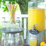 A rustic farmhouse-style lemonade station on a wooden porch featuring a large glass dispenser filled with MiMi’s Lemonade made from Country Time and orange juice. Surrounded by mason jars, striped paper straws, and fresh lemons, the setup evokes a cozy summer gathering in the country.