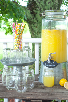 A rustic farmhouse-style lemonade station on a wooden porch featuring a large glass dispenser filled with MiMi’s Lemonade made from Country Time and orange juice. Surrounded by mason jars, striped paper straws, and fresh lemons, the setup evokes a cozy summer gathering in the country.