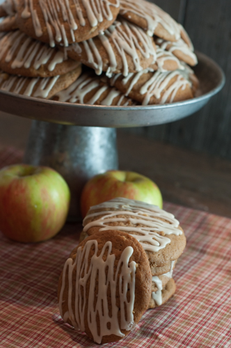 Large iced apple cider cookies stacked in front of a pedestal piled with cookies and fresh apples.