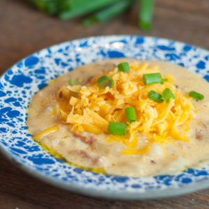 A bowl of creamy Crock Pot Loaded Potato Soup topped with shredded cheese and green onions, served on a rustic blue and white enamel plate. In the background, a crock pot, cheese grater, green onions, and a block of cheddar are visible, adding to the cozy kitchen atmosphere.