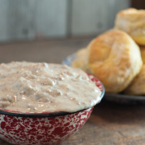 A red and white enamel bowl full of creamy homemade sausage gravy with a plate of flaky biscuits on a wooden table.