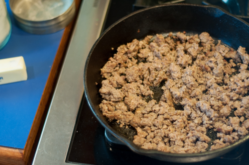 A cast iron skillet with crumbled browned pork sausage on a gas stove.