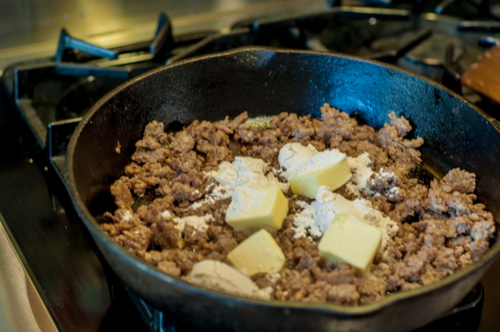 A cast iron skillet on a gas stovetop with browned crumbled pork sauasage with butter cubes and flour to make a roux for Homemade Sausage Gravy