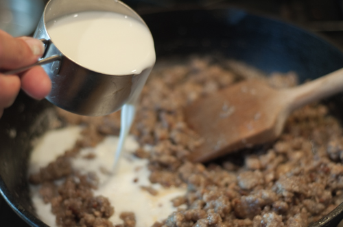 A cast iron skillet with crumbled cooked pork sausage  with milk being added and a wooden spoon for incorporating to make Sausage Gravy.