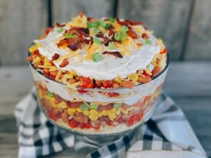 A vibrant, layered cornbread salad served in a glass trifle dish, featuring colorful layers of crumbled cornbread, pinto beans, corn, tomatoes, bell peppers, green onions, shredded cheese, crispy bacon, and creamy ranch dressing, garnished with green onions and bacon crumbles on top.