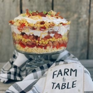 A vibrant, layered cornbread salad served in a glass trifle dish, featuring colorful layers of crumbled cornbread, pinto beans, corn, tomatoes, bell peppers, green onions, shredded cheese, crispy bacon, and creamy ranch dressing, garnished with green onions and bacon crumbles on top.