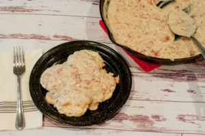 Plate of creamy dried beef gravy served over feshly baked biscuits, next to a skillet filled with the rich, savory gravy on a white barnwood table.