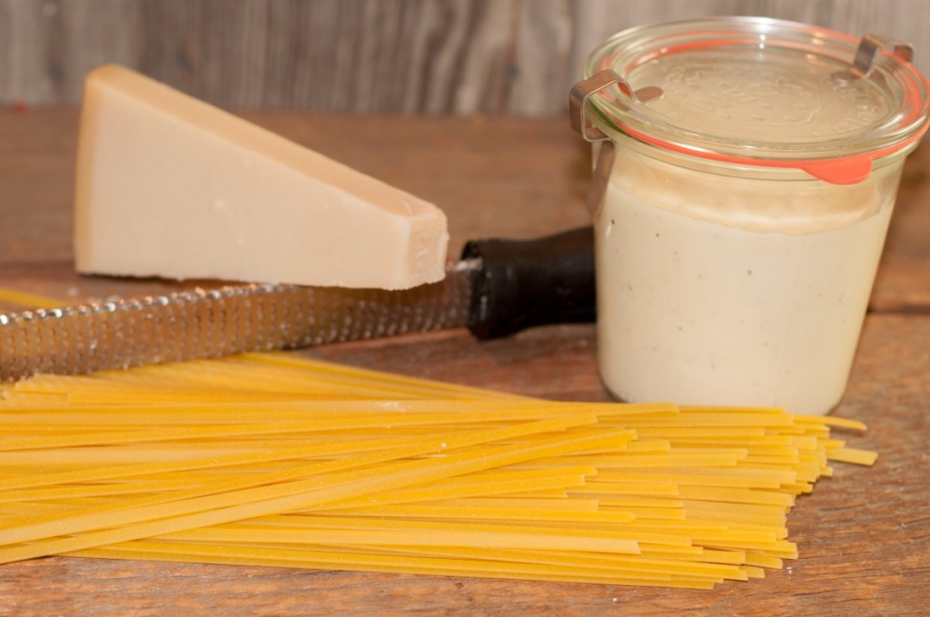Homemade Alfredo sauce stored in a glass jar next to uncooked pasta and Parmesan cheese