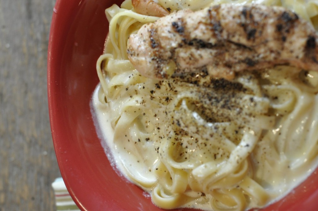 Close-up of creamy fettuccine Alfredo topped with grilled chicken and cracked black pepper in a red bowl