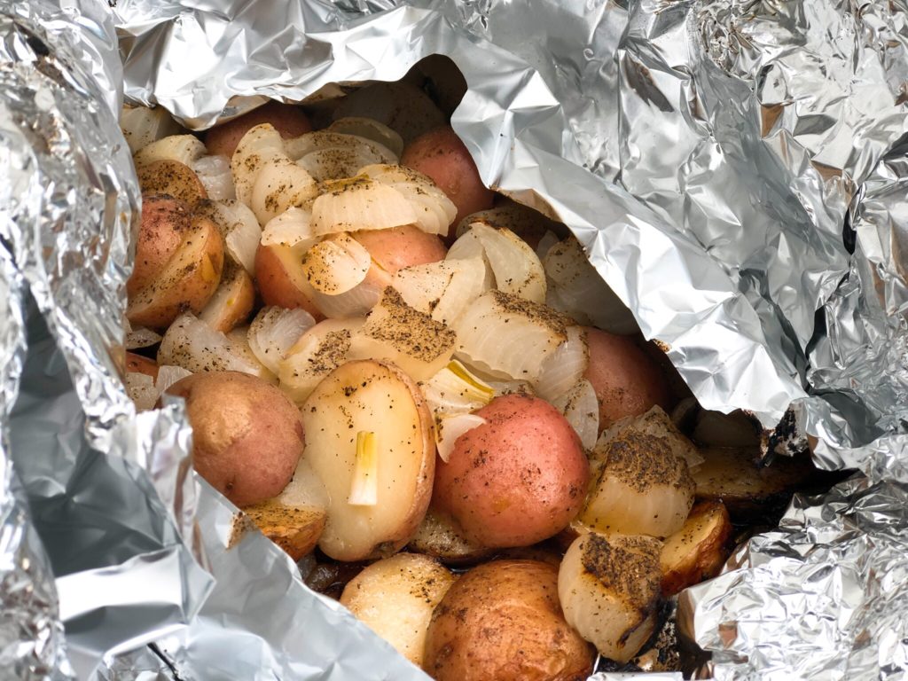 Foil packet filled with halved red potatoes and soft grilled onions, coated with melted butter and pepper, ready to serve