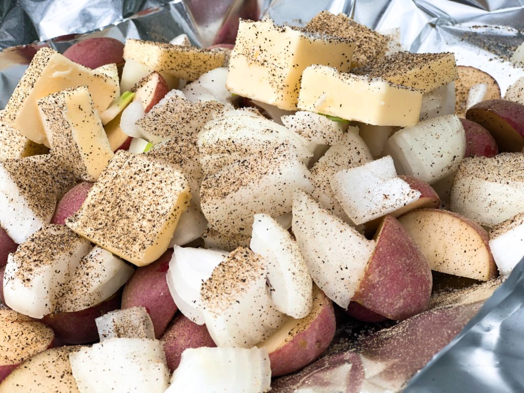 Raw foil packet ingredients including sliced butter, red potatoes, and chopped onions heavily sprinkled with pepper.