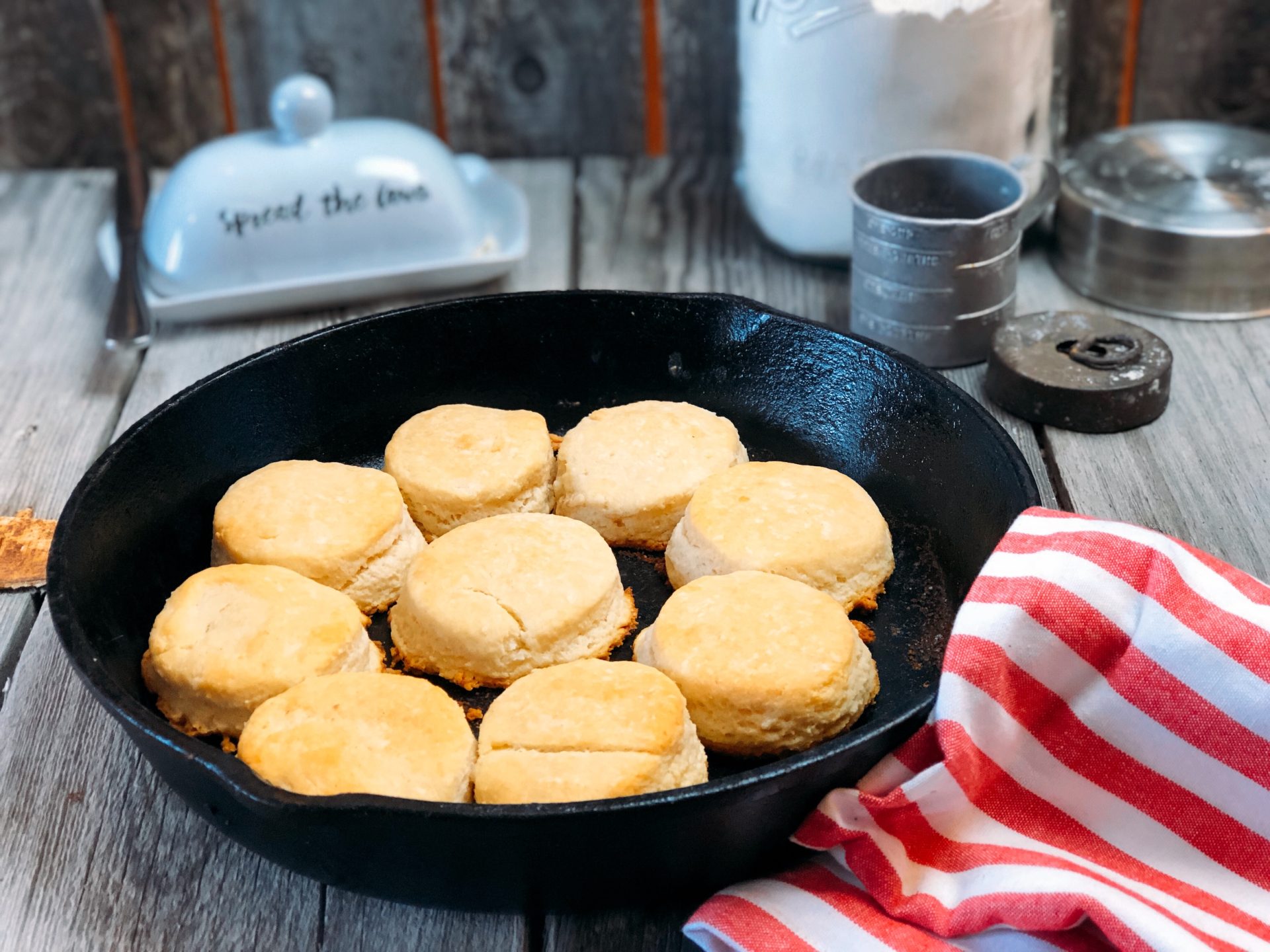 Golden brown buttermilk biscuits baked in a cast iron skillet, surrounded by baking tools and ingredients, with a red and white striped cloth for a rustic kitchen setting.