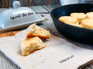 Freshly baked buttermilk biscuit split open to reveal its fluffy interior, set on a cloth with the word 'savor,' alongside a butter dish with 'spread the love' written on it.