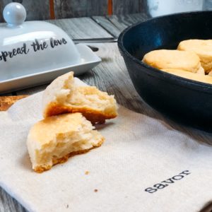 Freshly baked buttermilk biscuit split open to reveal its fluffy interior, set on a cloth with the word 'savor,' alongside a butter dish with 'spread the love' written on it.