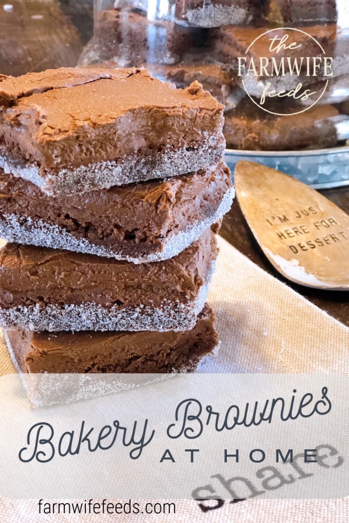 Stack of freshly baked brownies sprinkled with sugar on a rustic kitchen setting, with text overlay 'Bakery Brownies at Home' and 'The Farmwife Feeds' logo, promoting homemade dessert recipes at farmwifefeeds.com