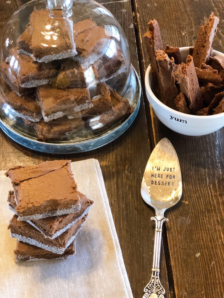 Stack of homemade brownies with sugar edges on a wooden table, featuring decorative kitchen items like a glass dome full of brownies and a dessert spoon with the inscription 'I'm just here for dessert', food and lifestyle blogging at farmwifefeeds.com