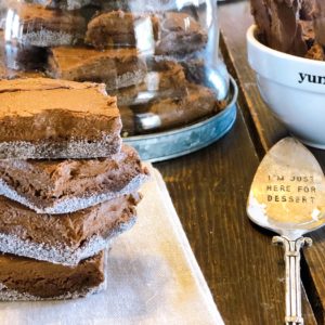 Stack of homemade brownies with sugar edges on a wooden table, featuring decorative kitchen items like a glass dome full of brownies and a dessert spoon with the inscription 'I'm just here for dessert', food and lifestyle blogging at farmwifefeeds.com
