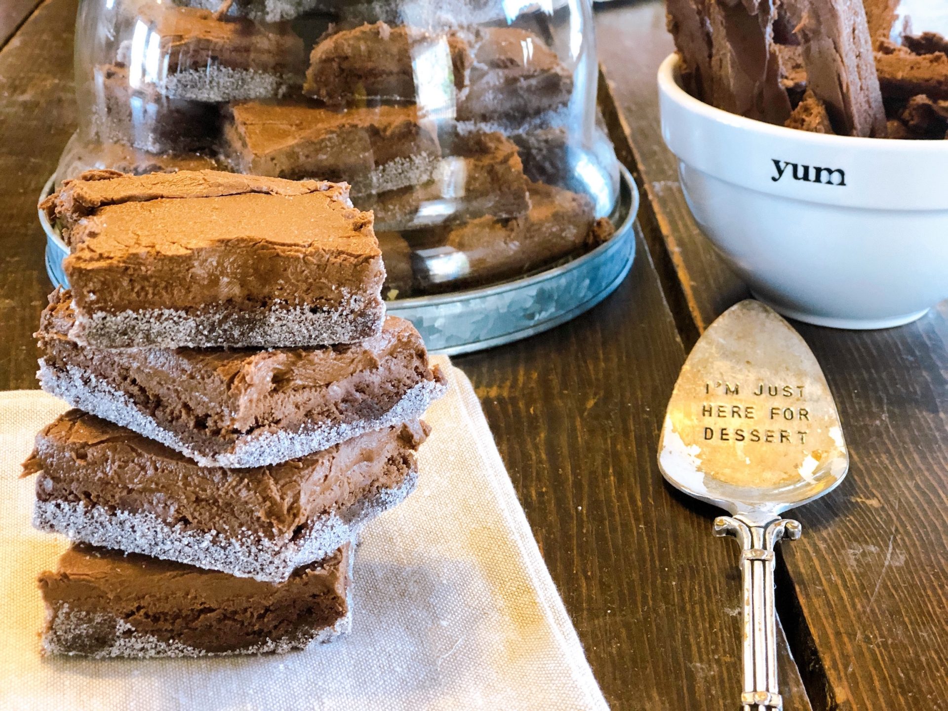 Stack of homemade brownies with sugar edges on a wooden table, featuring decorative kitchen items like a glass dome full of brownies and a dessert spoon with the inscription 'I'm just here for dessert', food and lifestyle blogging at farmwifefeeds.com