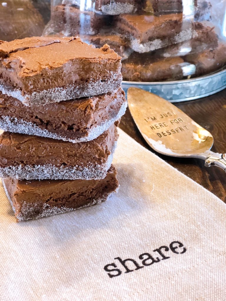 Stack of homemade brownies with sugar edges on a wooden table, featuring decorative kitchen items like a glass dome full of brownies and a dessert spoon with the inscription 'I'm just here for dessert', food and lifestyle blogging at farmwifefeeds.com