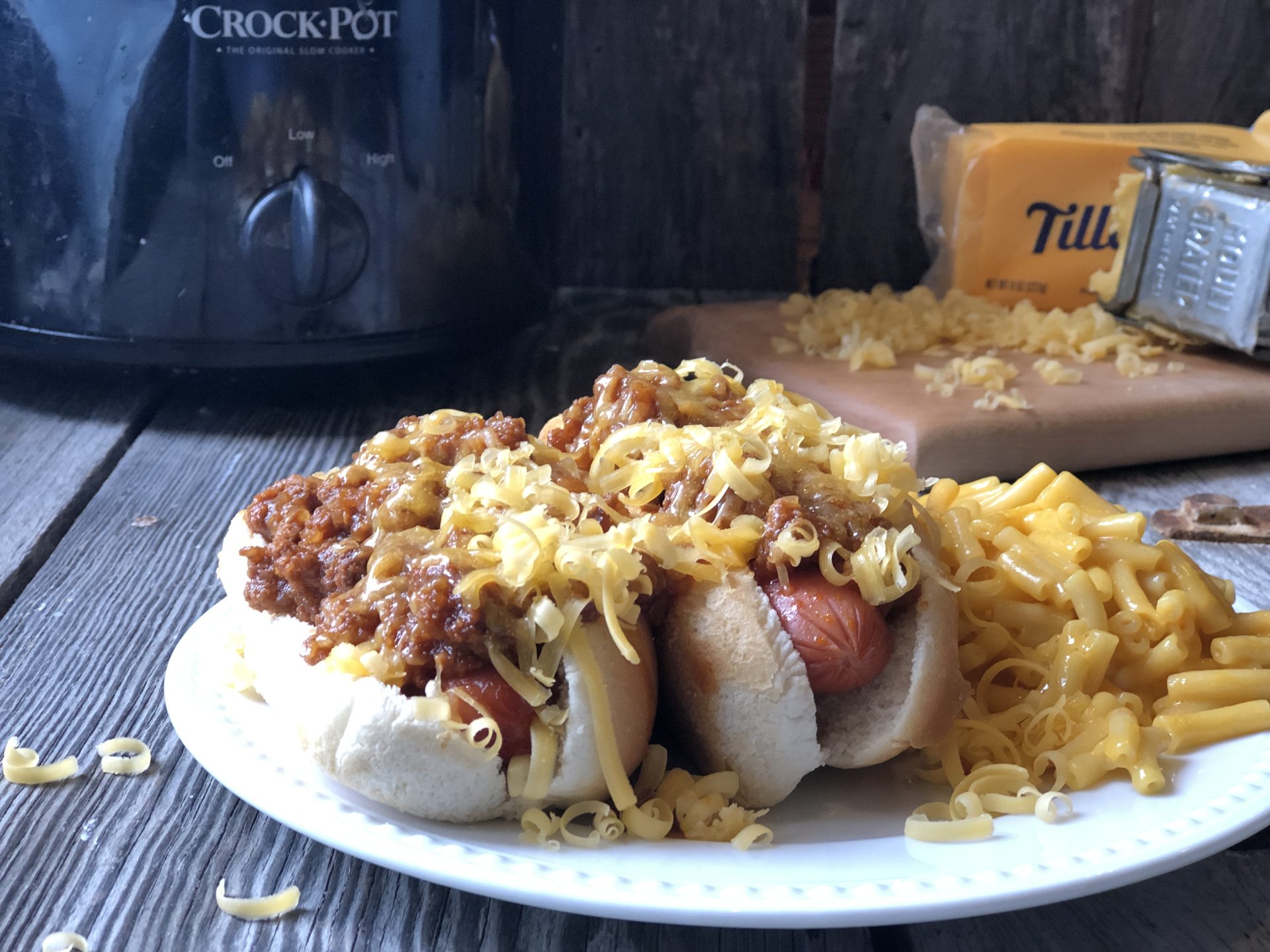 Plate of hot dogs topped with traditional coney dog sauce and shredded cheese, served with macaroni and cheese, with a Crock-Pot, block of cheddar, and a cheese grater on a rustic wood table in the background.