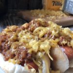 Plate of hot dogs topped with traditional coney dog sauce and shredded cheese, served with macaroni and cheese, with a Crock-Pot, block of cheddar, and a cheese grater on a rustic wood table in the background.
