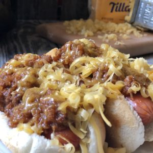 Plate of hot dogs topped with traditional coney dog sauce and shredded cheese, served with macaroni and cheese, with a Crock-Pot, block of cheddar, and a cheese grater on a rustic wood table in the background.