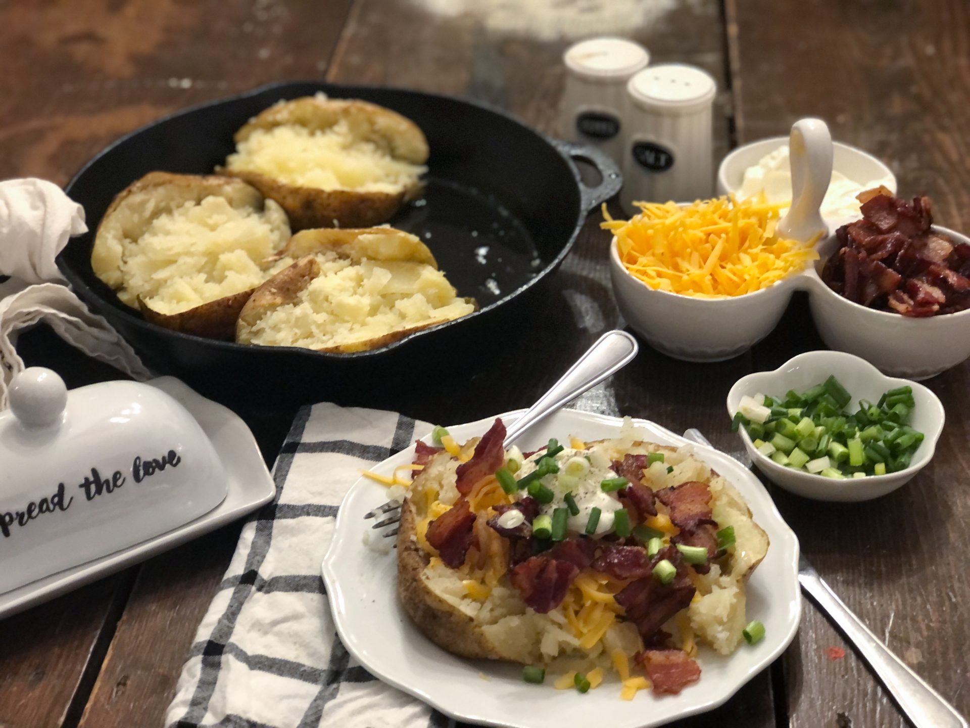 cast iron skillet with baked potatoes plate with baked potatoe with garnish bacon green onions cheese on table