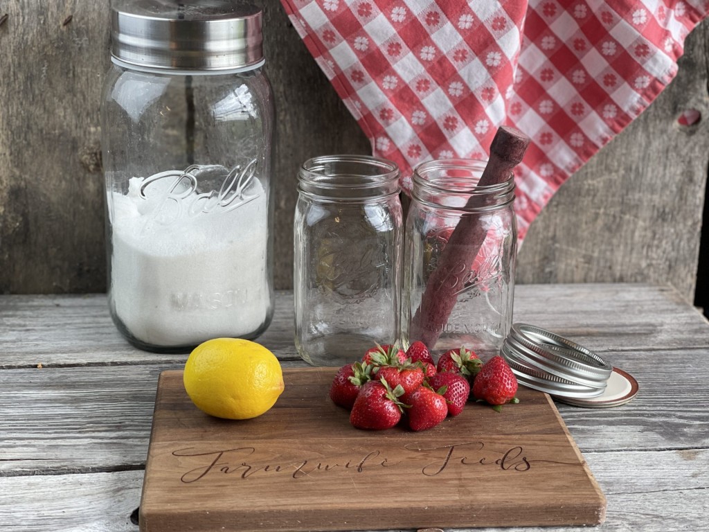 mason jar of granulated sugar 2 smaller mason jars a muddler and lemon and strawberries on a wooden board