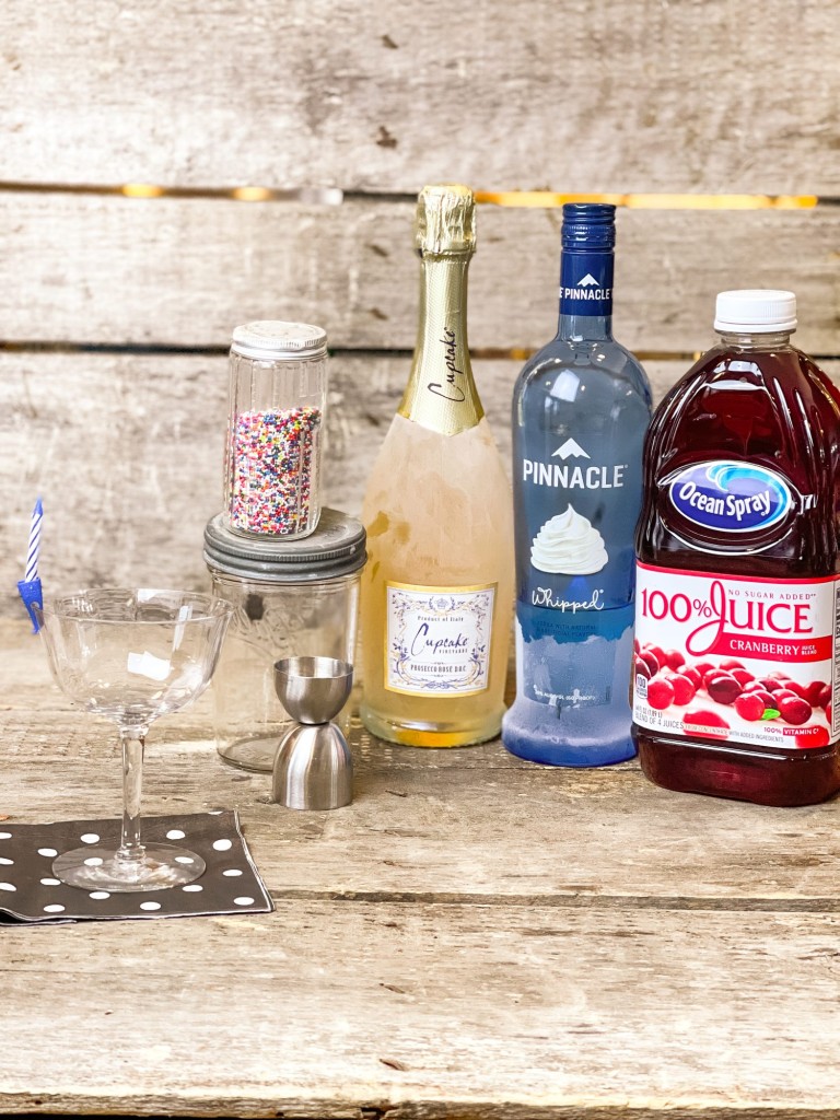 Cocktail glass with a birthday candle on the rim on a black and white polka dot napkin next to a glass jar of rainbow sprinkles or non-perils