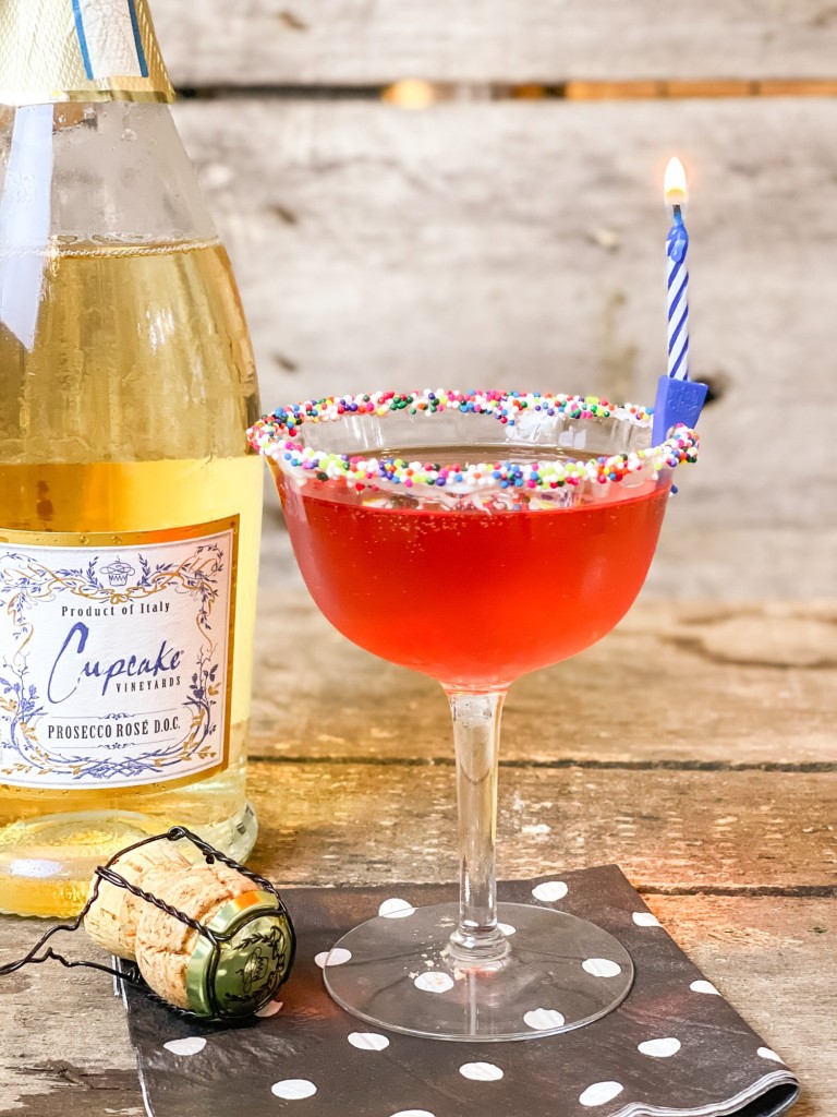 bottle of Cupcake Prosecco, martini glass rimmed in colorful sprinkles on a black white polka dotted napkin