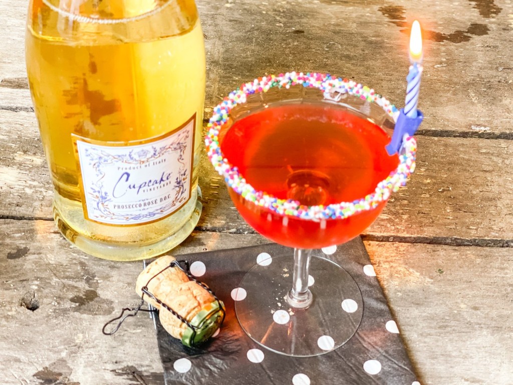 bottle of prosseco black white polka dot napking cocktail glass rimmed in sprinkles and a birthday candle with a red pink drink