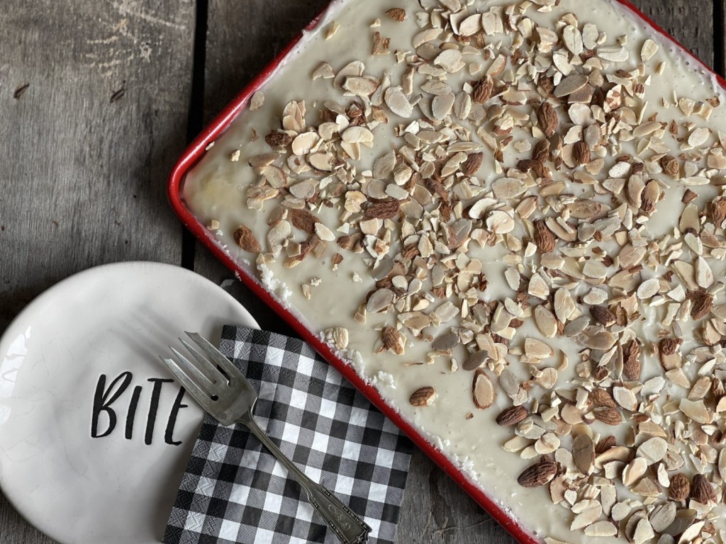 White Texas Almond Sheet cake in a red pan covered in white icing with slivered almonds on top on a wood grain background