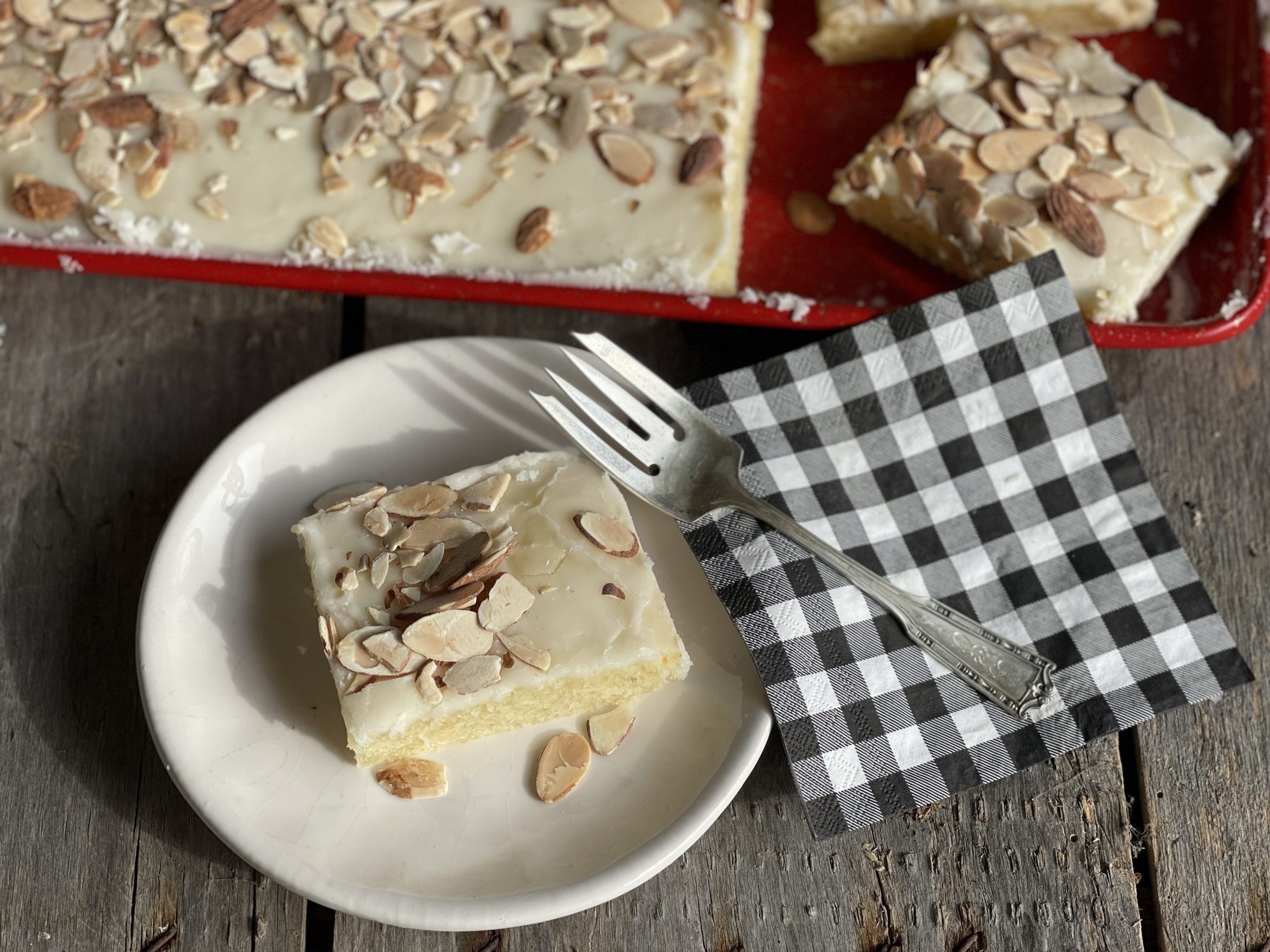 Slice of White Almond Texas Sheet cake on a plate with red serving pan of cake in the back ground