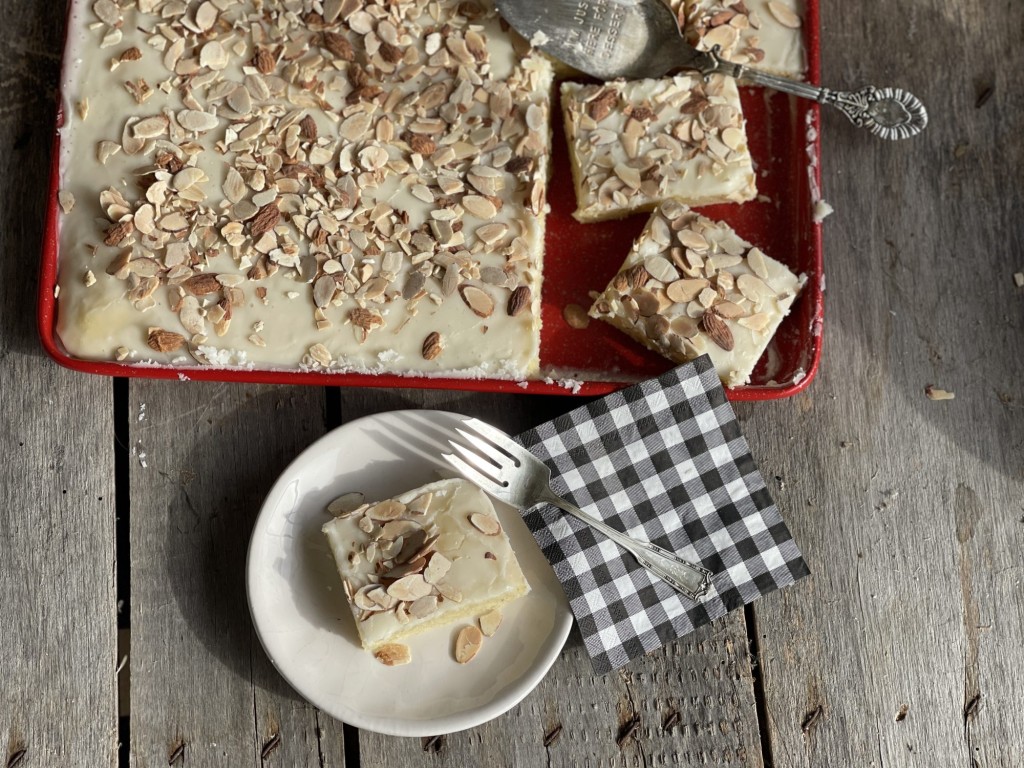 Slice of White Almond Texas Sheet cake on a plate with red serving pan of cake in the back ground