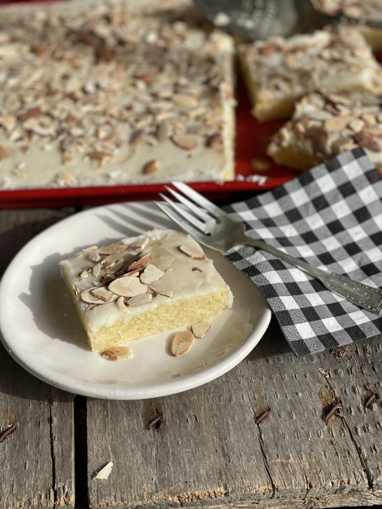 Slice of White Almond Texas Sheet cake on a plate with red serving pan of cake in the back ground
