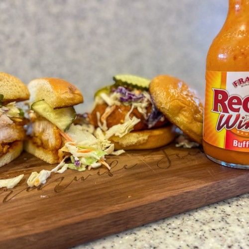Two Air Fryer Crispy Buffalo Chicken Sandwiches with coleslaw and pickles on a toasted brioche bun on a wooden cutting board with a bottle of Franks Red Hot Wind Sauce