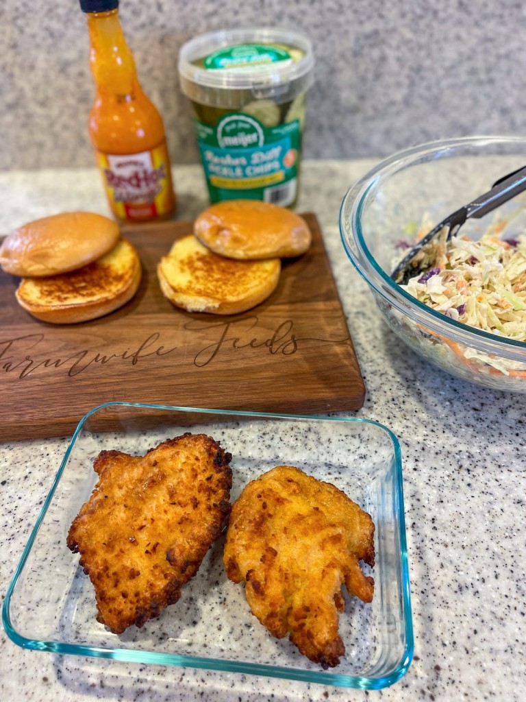 Crispy Buffalo Chicken sandwich assembly line- two toasted buns two cripsy chicken breasts coated in Franks red hot wing sauce and a bowl of coleslaw