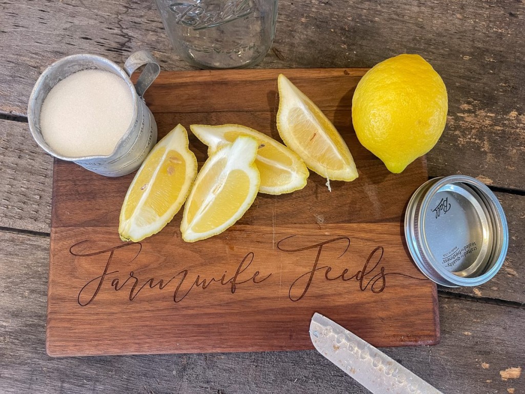 A whole lemon and a quartered lemon with a measuring cup of sugar on a wooden cutting board