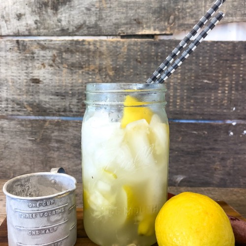 A refreshing lemon shake-up served in a mason jar, accompanied by a vintage measuring cup and a fresh lemon on a rustic wooden cutting board. The drink is chilled with ice cubes, garnished with lemon slices, and ready to sip with striped paper straws, conveying a perfect summer refreshment vibe. The backdrop features a weathered wooden surface, enhancing the farm-style aesthetic.