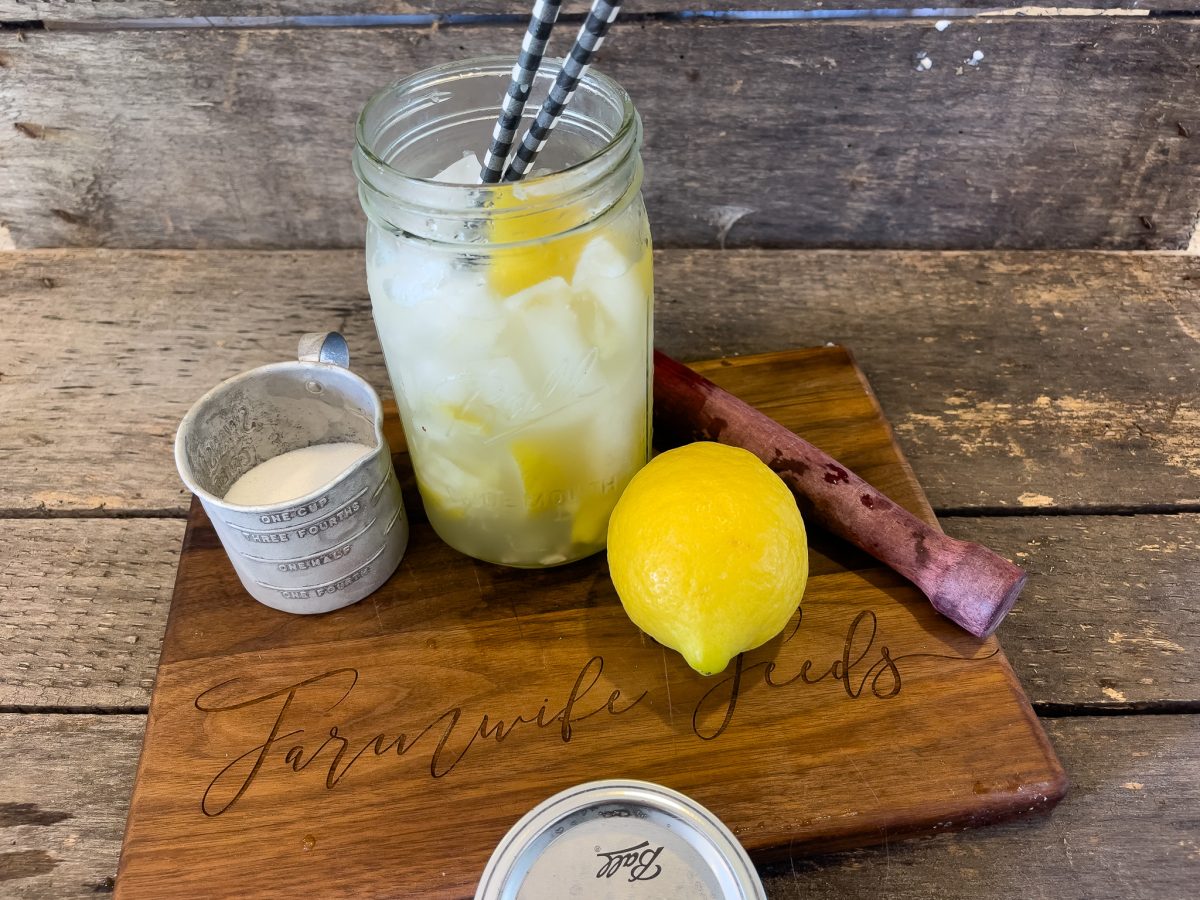 A refreshing lemon shake-up served in a mason jar, accompanied by a vintage measuring cup and a fresh lemon on a rustic wooden cutting board. The drink is chilled with ice cubes, garnished with lemon slices, and ready to sip with striped paper straws, conveying a perfect summer refreshment vibe. The backdrop features a weathered wooden surface, enhancing the farm-style aesthetic.
