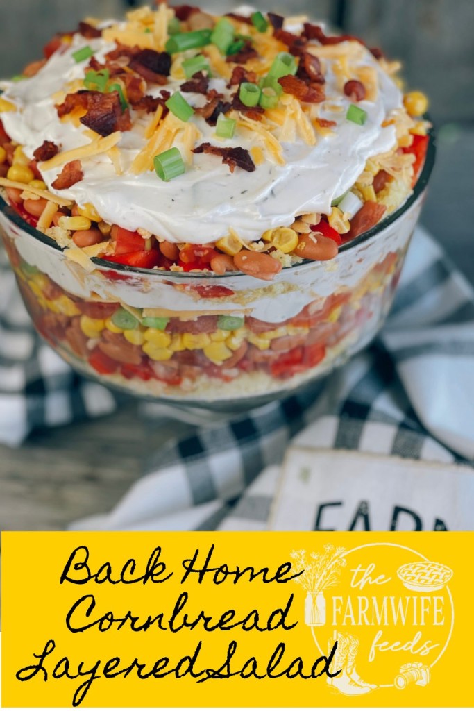 A vibrant, layered cornbread salad served in a glass trifle dish, featuring colorful layers of crumbled cornbread, pinto beans, corn, tomatoes, bell peppers, green onions, shredded cheese, crispy bacon, and creamy ranch dressing, garnished with green onions and bacon crumbles on top.