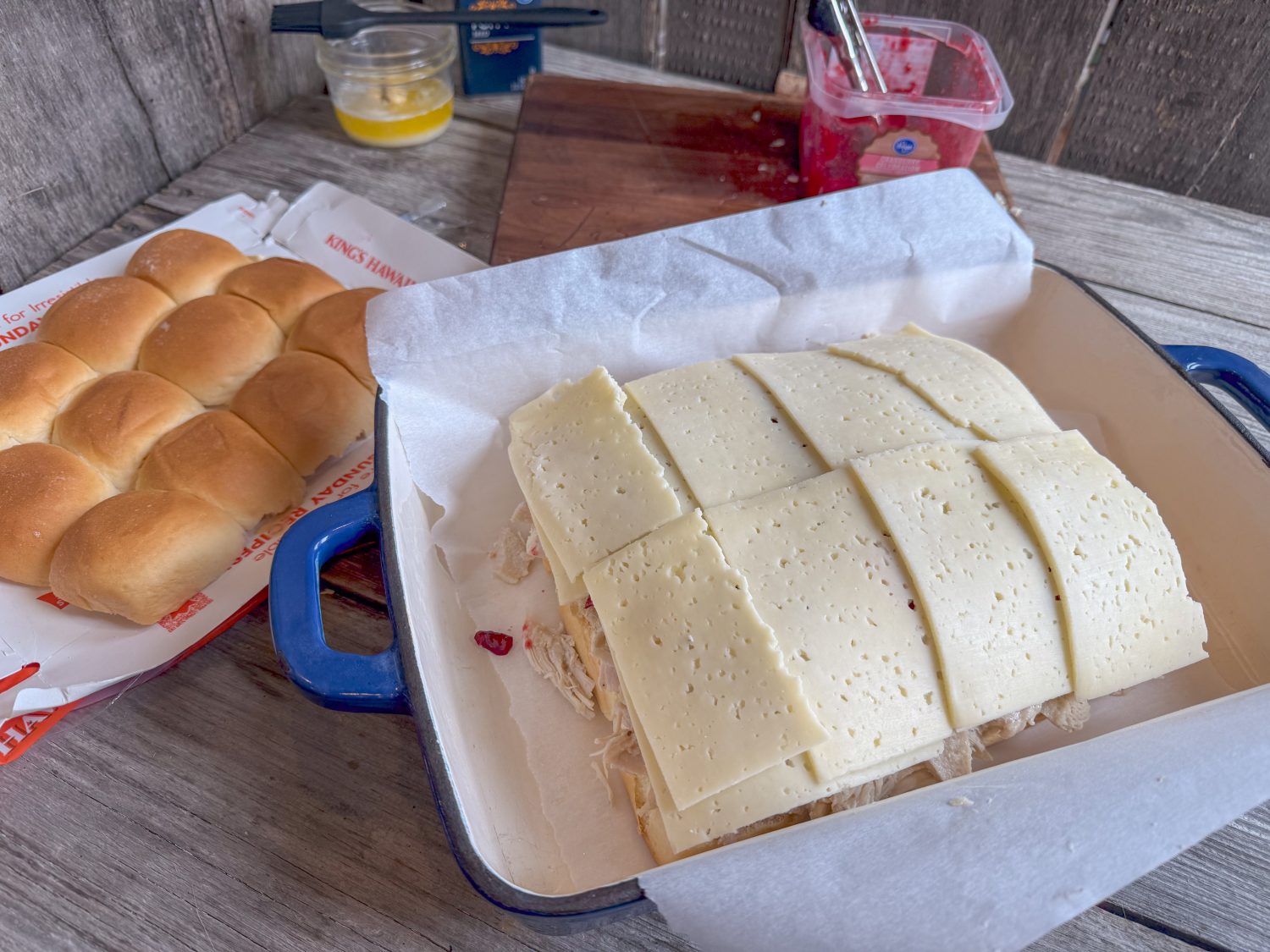 Slices of Havarti cheese layered over turkey and cranberry sauce in a baking dish, with Hawaiian rolls and ingredients nearby.