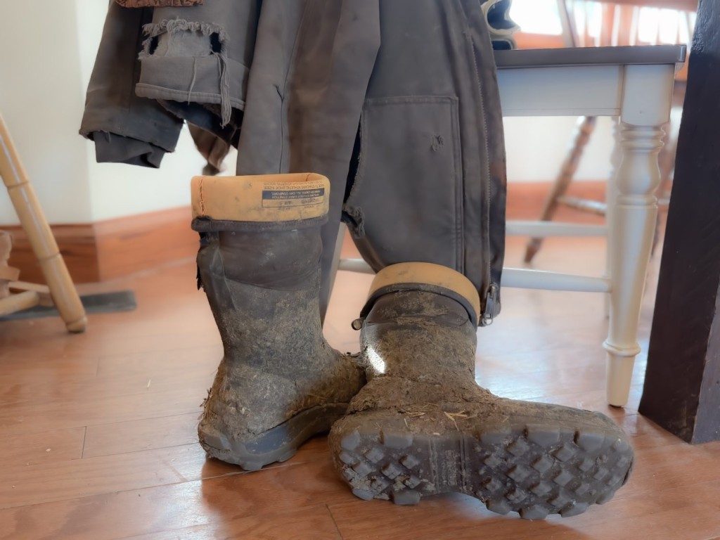 Muddy boots and a torn Carhartt jacket sitting my a kitchen chair, showing the exhaustion and chaos of farm mom life during harvest.