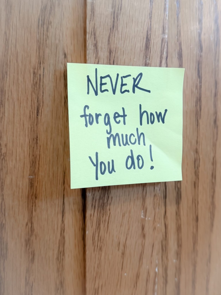 Yellow Post-it note on a wooden kitchen cabinet with the handwritten message "Never forget how much you do," a reminder of strength during hard times.