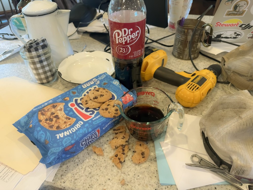 Measuring cup filled with flat Dr Pepper and a stack of broken chocolate chip cookies on a messy kitchen counter, symbolizing farm mom burnout.