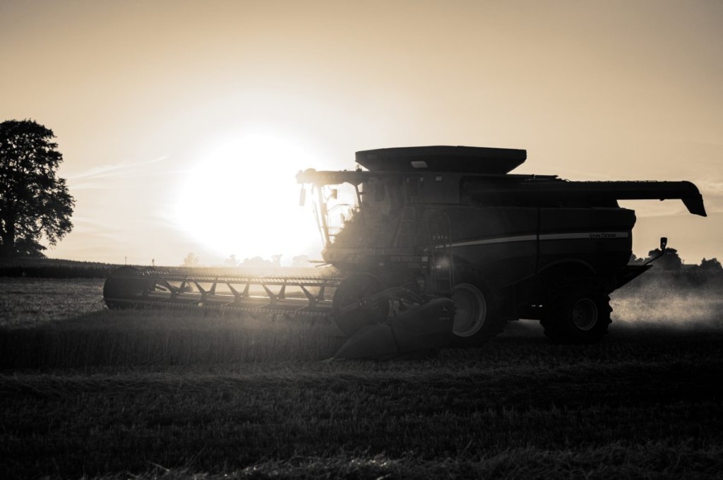 A JohnDeere combine silhouetted against a glowing sunset druing harvest, with dust rising from the field in a peaceful end-of-day scene.