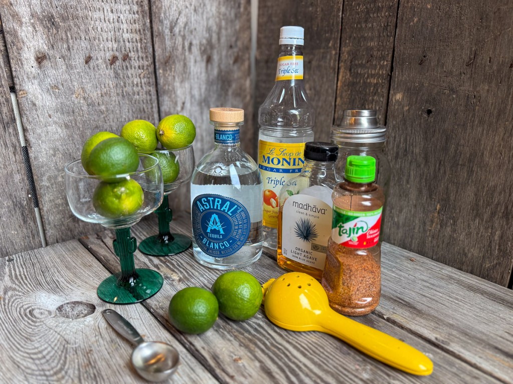 Skinny margarita ingredients on a rustic wood surface including fresh limes, Astral tequila, Monin sugar free triple sec syrup, Madhava organic agave, Tajín seasoning, and margarita glasses.